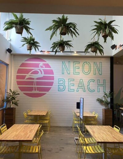 A trendy indoor dining area with a "neon beach" sign on the wall, surrounded by hanging potted plants and wooden tables with yellow chairs.