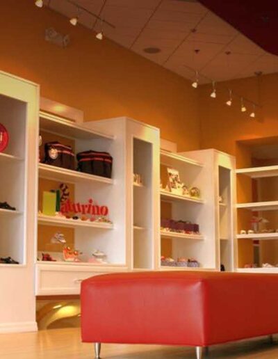 A brightly lit shoe store interior with white shelves displaying various footwear and a red bench for seating.