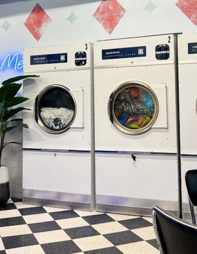 A retro-styled laundromat with a neon sign that reads "talk dirty to me," featuring a row of washing machines, one being out of order.
