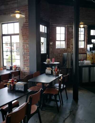 Interior of a casual dining area with wooden tables and chairs, exposed brick walls, and a beverage station in the corner.