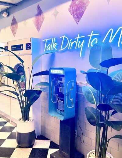 A neon-lit laundromat with a retro payphone and decorative plants.