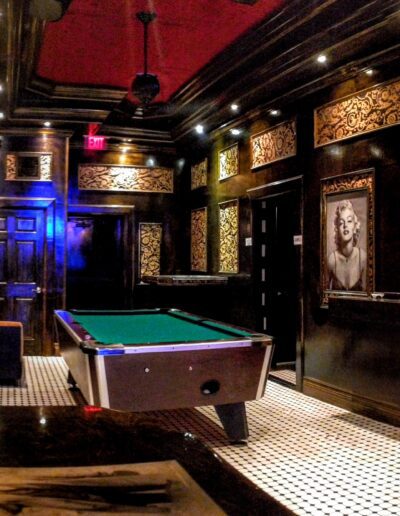 An ornately decorated room with a pool table, bar stools, and art-adorned walls, illuminated with dim, ambient lighting.