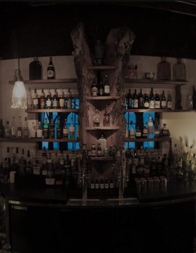 Panoramic view of a dimly lit bar with an array of bottles on shelves and a wooden beam ceiling.